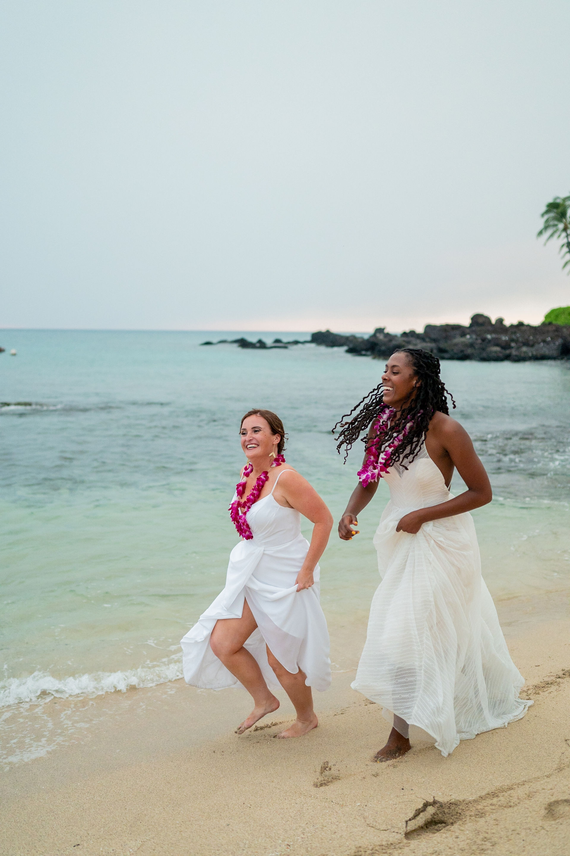 Two people in white dresses and pink leis run on Kona beach near ocean waves and rocky shoreline.