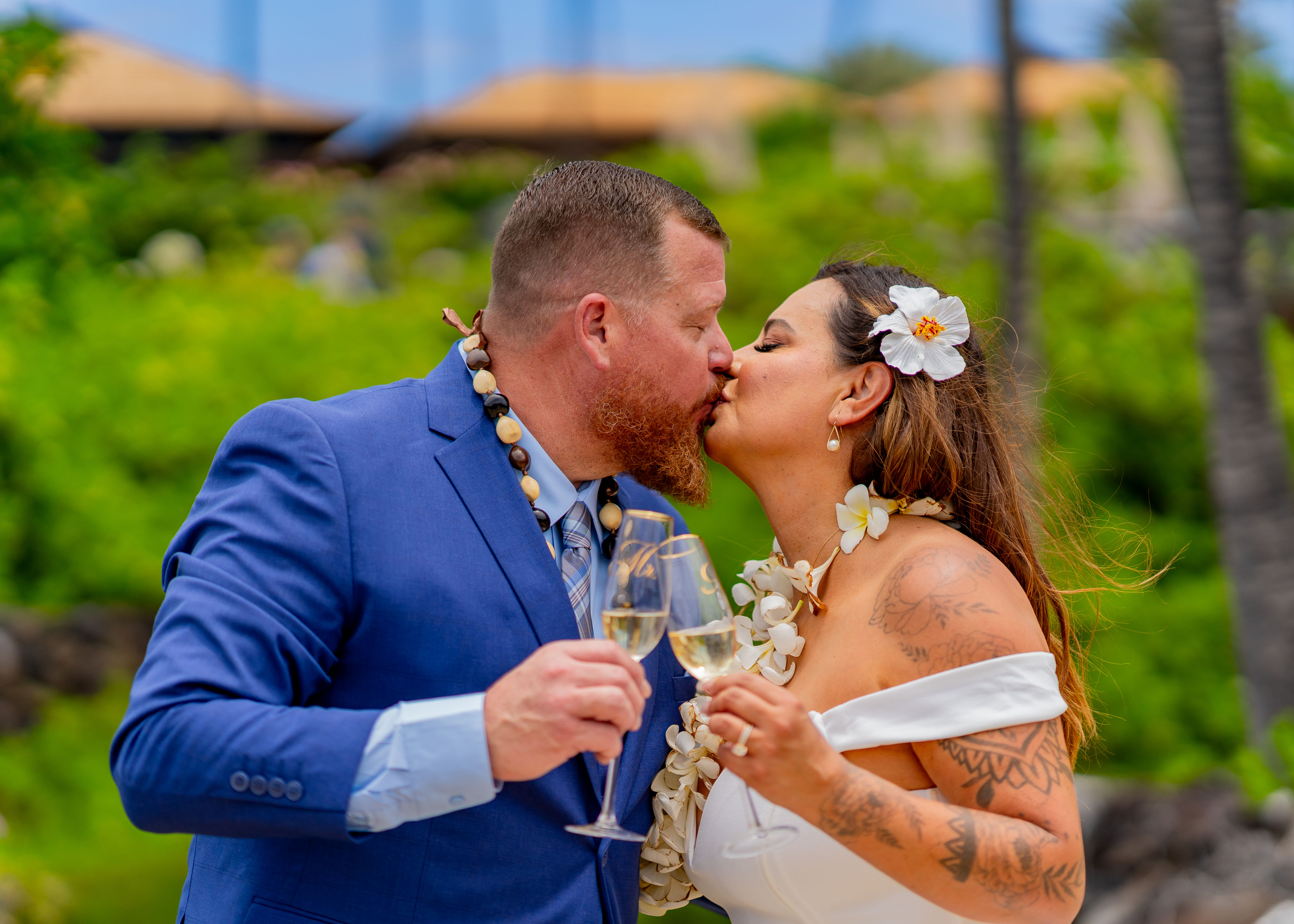 “Couple kisses with champagne in hand, floral attire and greenery near Kona Wedding Officiant.”