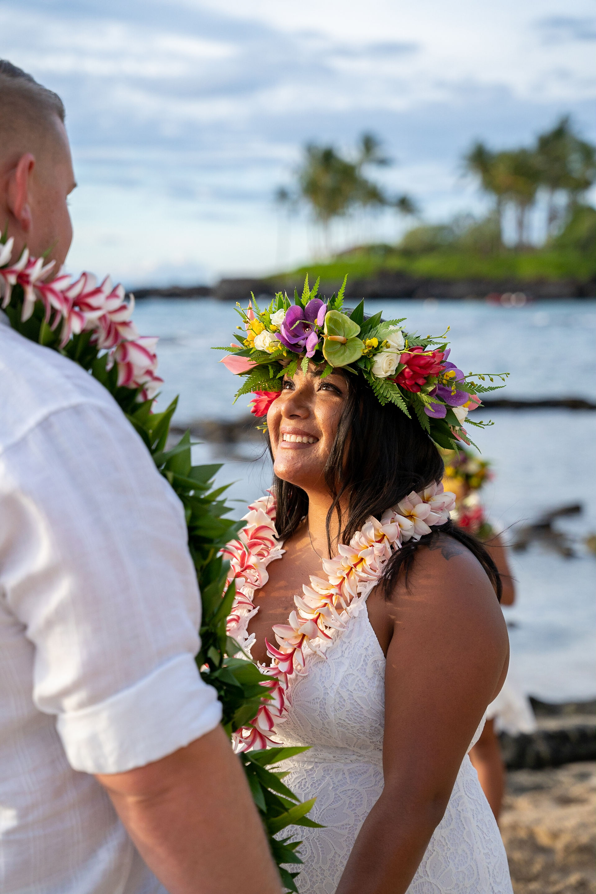 Couple in leis and floral crowns shares joyful gaze on Kona beach with ocean, rocks, and palm trees.