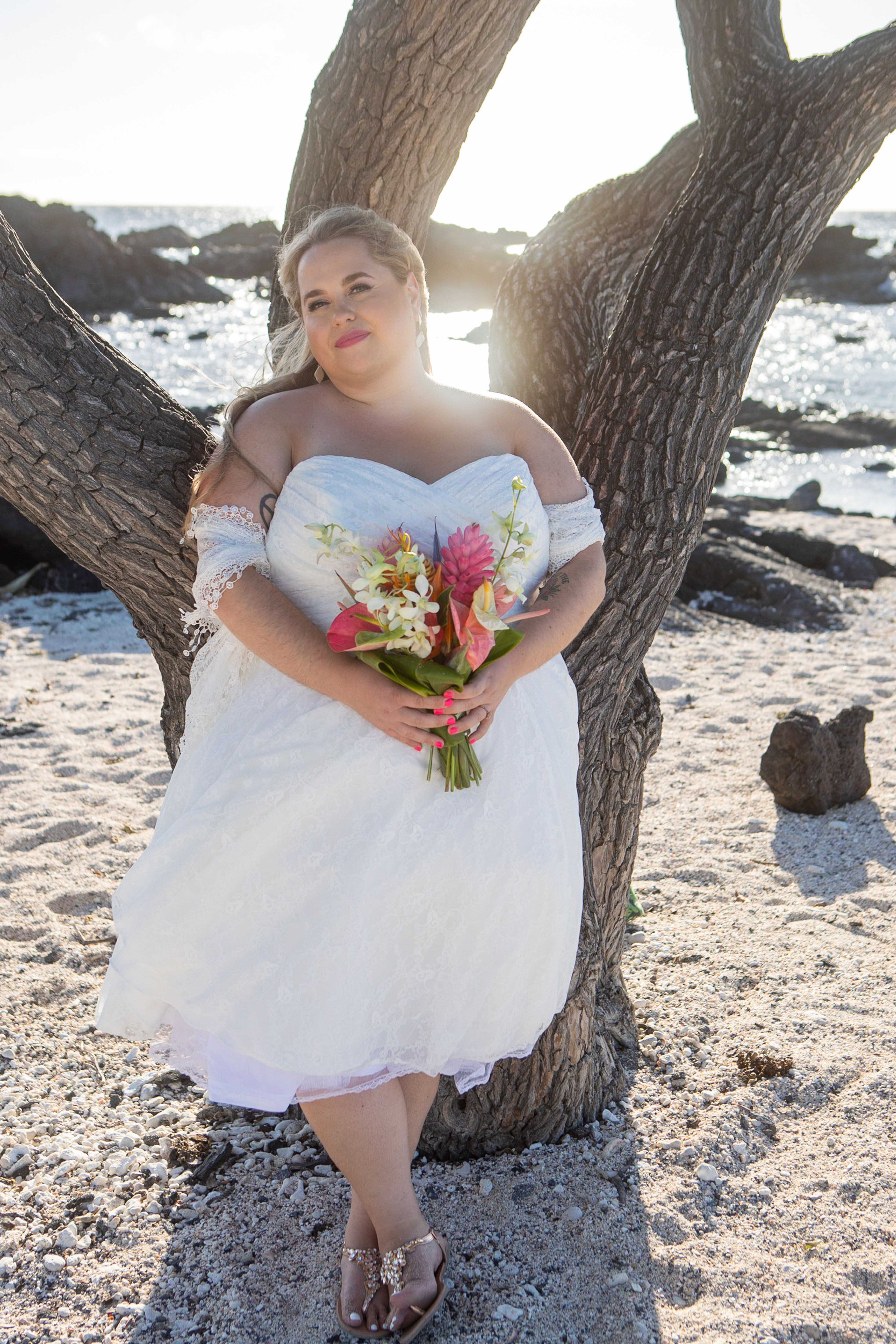 Person in white wedding dress stands on Kona’s sandy shore, bouquet in hand, backlit by sunlight through tree branches with o