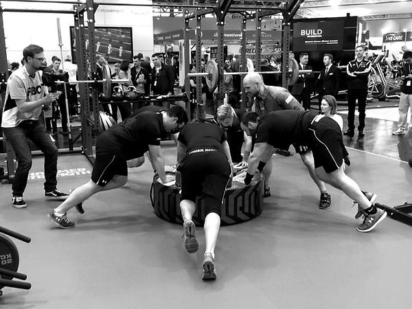 Eine Gruppe von fünf Menschen führt eine Sportübung aus. Sie stützen sich unter der Anleitung von Marc Rohde mit den Händen auf einem Reifen auf. Im Hintergrund stehen Sportgeräte in einem Messe Umfeld.