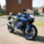 Blue Yamaha R125 motorcycle parked on a brick driveway with a sign saying "Sell Your 125cc Motorbike today." Sunny suburban background.