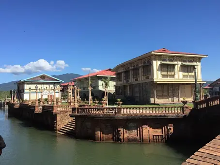A row of traditional Filipino homes with capiz windows, elevated stone foundations, and red-tiled roofs beside a man-made canal under a clear blue sky.