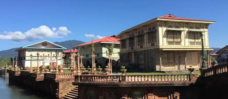 A row of traditional Filipino homes with capiz windows, elevated stone foundations, and red-tiled roofs beside a man-made canal under a clear blue sky.