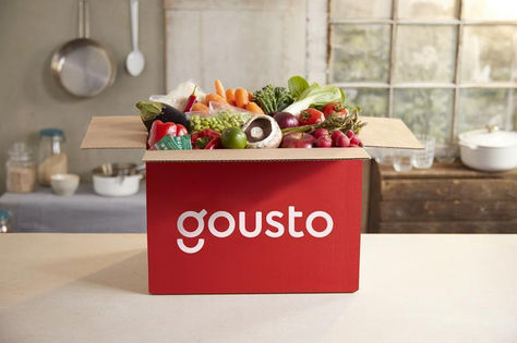 Gousto meal kit delivery box on a kitchen counter, filled with fresh vegetables and ingredients, illustrating convenient home cooking and reduced food waste.