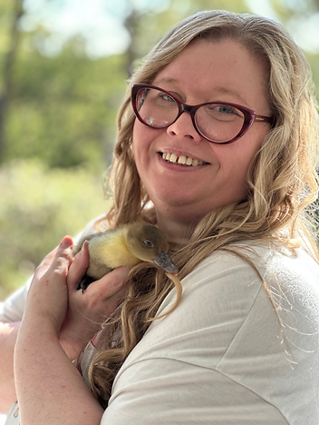 Kirsten, a white woman with curled blonde hair and pink glasses, in a white t shirt holding a yellow and brown duckling and smiling