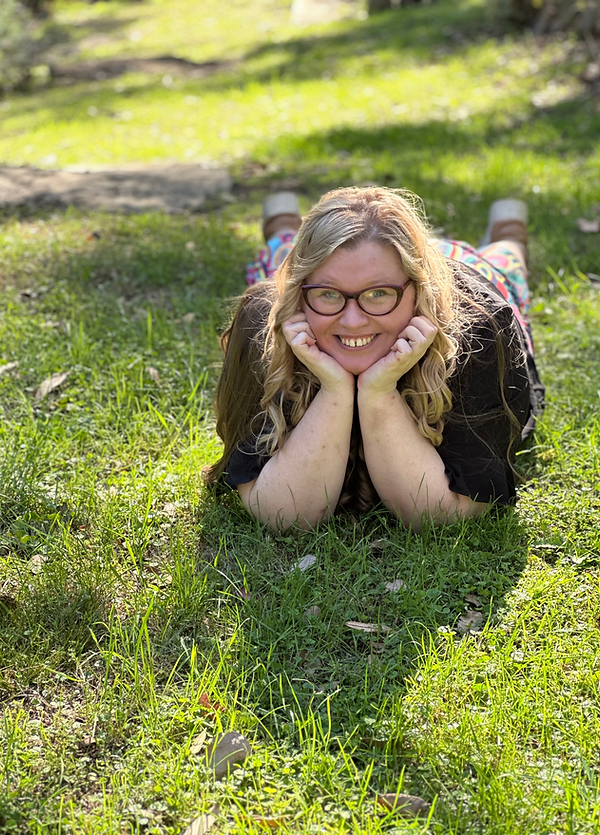 Picture of a white woman laid on the ground, surrounded by grass, with her head in her hands, smiling playfully