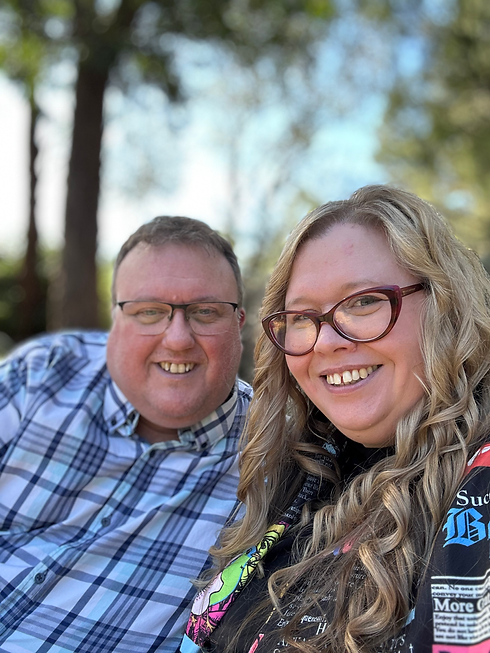 Kirsten and Tony sat together, looking at the camera and smiling. Tony is a white man, with glasses and in a blue and white checked shirt. Kirsten is a white woman with blonde curled hair and pink glasses