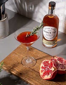 Pomegranate Manhattan cocktail on a cutting board, to the left of sliced pomegranate, which bottle fo Broken Top Rye in background.