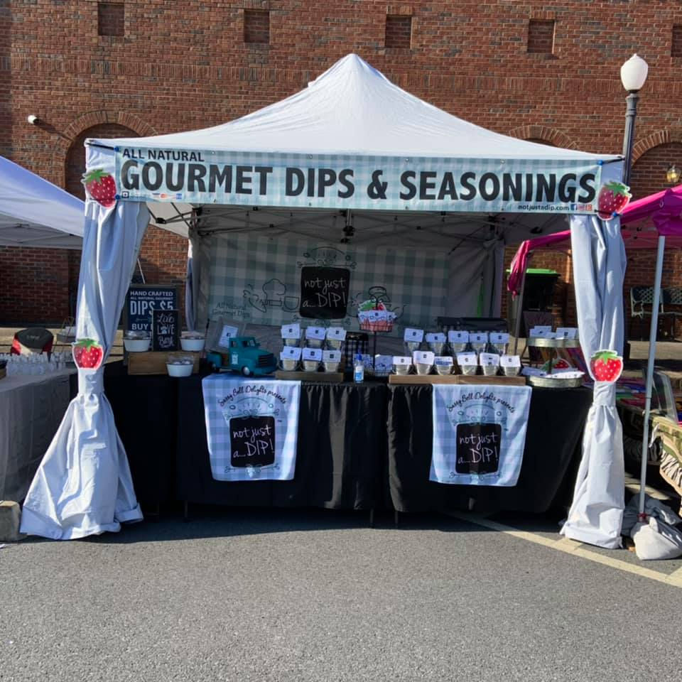 Outdoor market booth for All Natural Gourmet Dips & Seasonings, with jars, a checkered canopy, and strawberry accents.