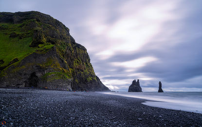 A Praia de Reynisfjara com sua areia negra, seus lendários três pilares de basalto (Reynisdeangar) e a Caverna Hálsanefshellir da erosão do basalto pelas ondas do mar, próxima da vila Vík í Mýrdal, Islândia