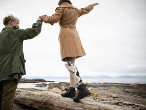 Person helping another person maintain balance while crossing a large driftwood log near the shoreline.