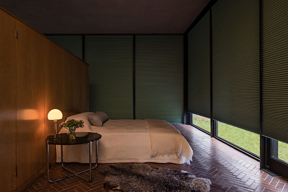 Cozy modern bedroom with a bed, glowing bedside lamp, flowers, Hunter Douglas honeycomb blinds with LightLock, wood paneling, brick floor, and shag rug.