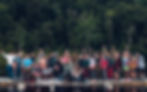 A group of people at Stargaze Festival pose on a dock by a forested lake, smiling and cheering. Text reads "Lesbian Nightlife." Casual attire, evening setting.
