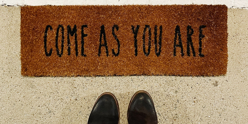 Brown doormat with "COME AS YOU ARE" in black letters, placed on a light concrete floor. Two black shoes visible at the bottom edge. Welcoming mood. Gene key 22 Grace.