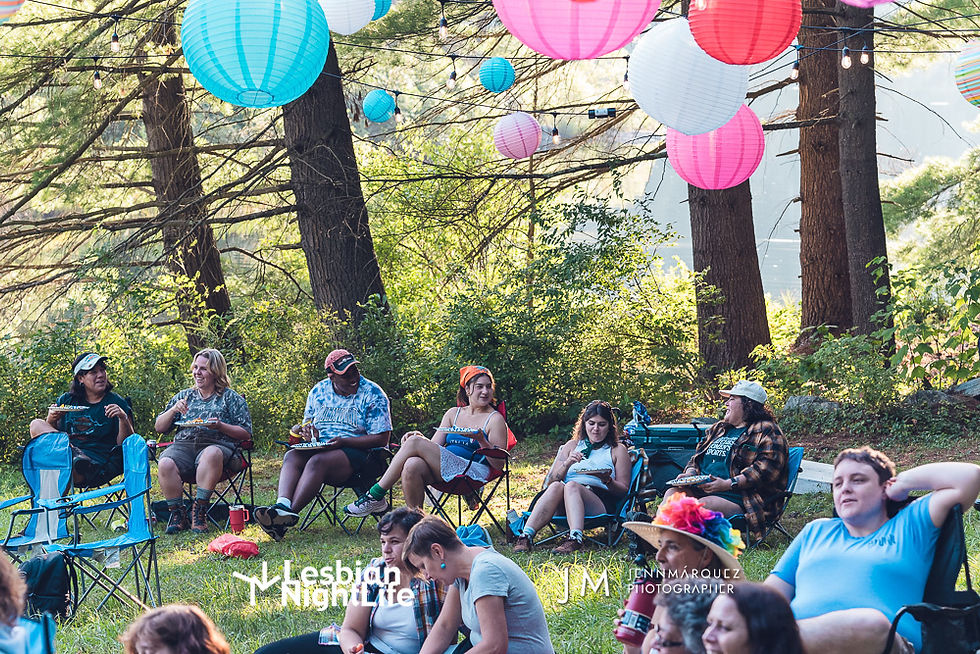 People relax in chairs under colorful lanterns in a forest setting. The scene feels festive and carefree. Visible text: Lesbian Night Life.