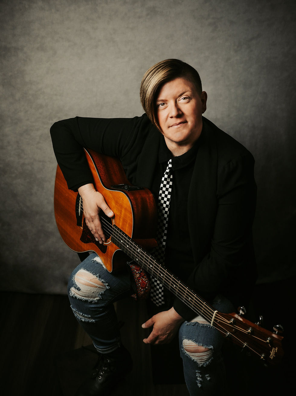 Andrea Paquin holds a guitar, wearing a black jacket and checkered tie. Sitting against a gray background, mood is calm.