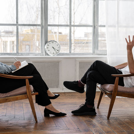 White female coach working with white male client. Each sitting in mid-century modern wood chairs in front of a wall of windows with sheer white curtains and a clock on the window sill in the middle of the photo.