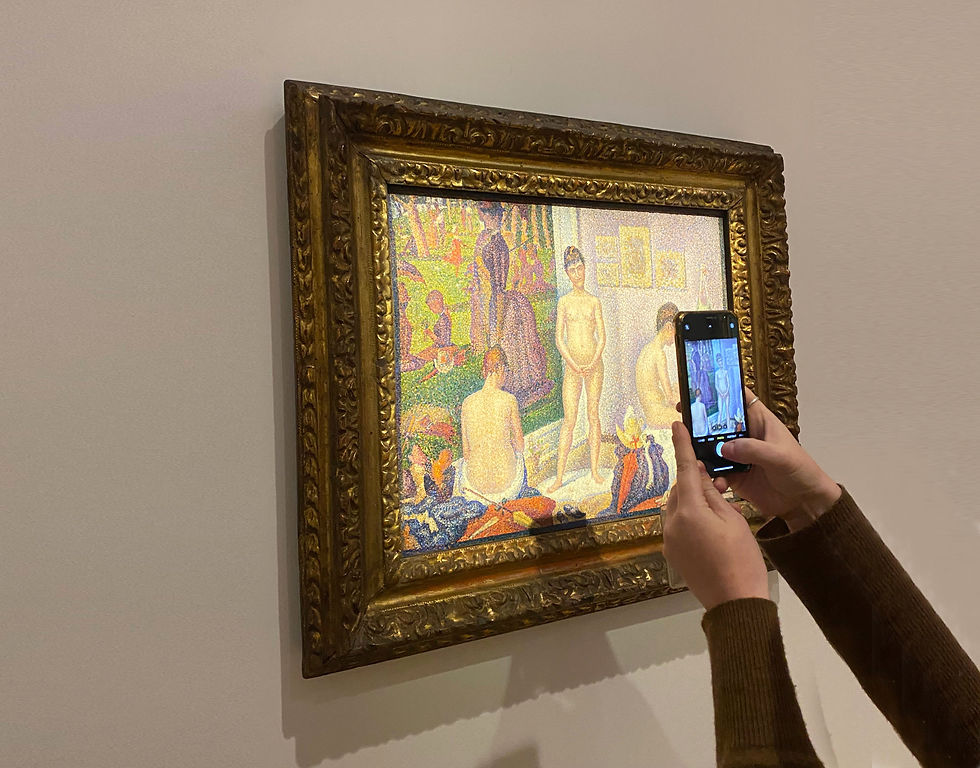 A person photographs a framed, colorful pointillism painting in a gallery. Hands hold a phone, capturing the artwork against a plain wall.