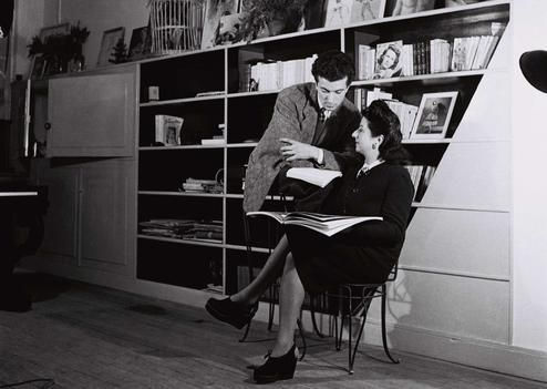Man and woman interact by a bookshelf; the man points at a book the woman holds. Black and white with a cozy, studious atmosphere.