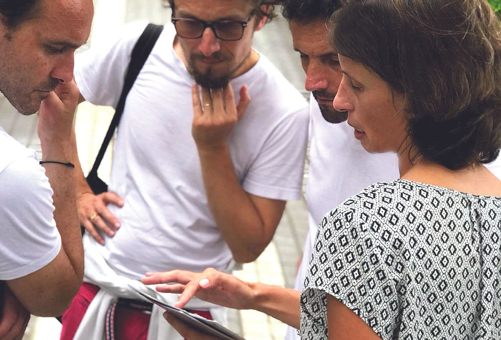 Four people gather, focused on a tablet. One points at it. They're wearing casual clothes. Background is blurred; mood is attentive.