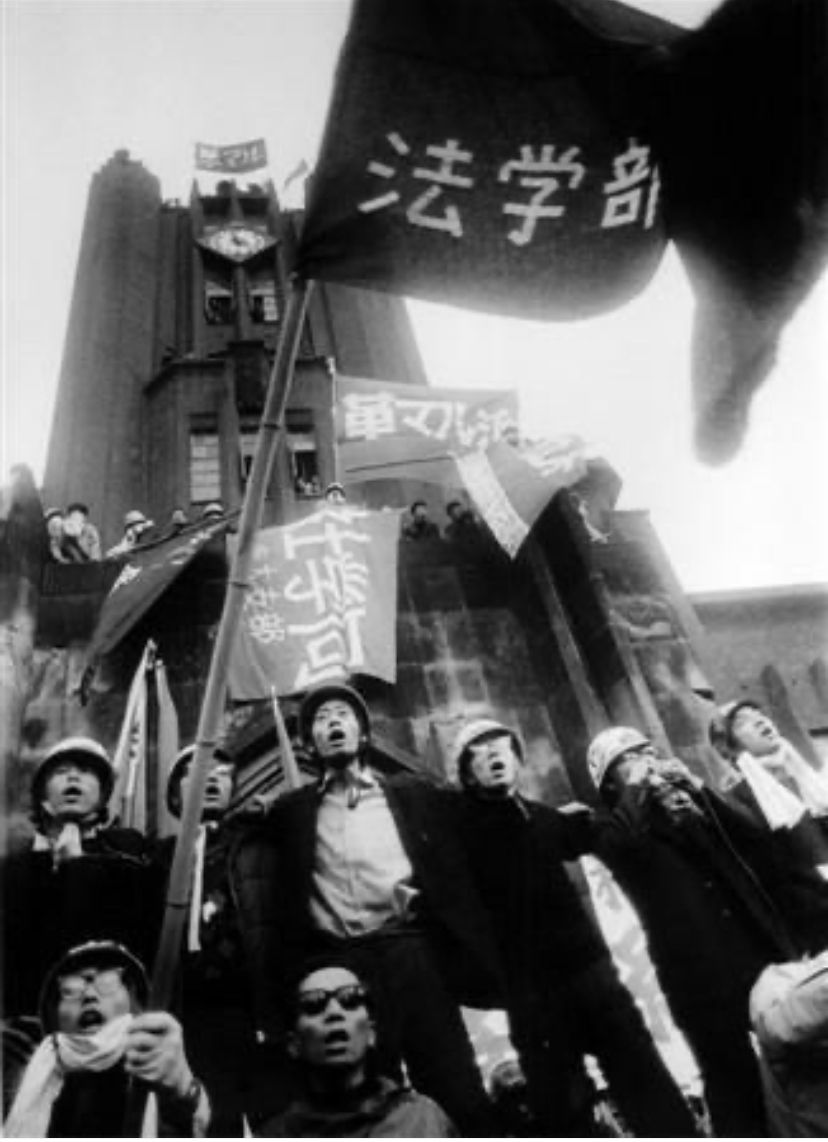 Protesters wearing helmets chant in front of a tall building holding flags with Japanese text, displaying determination and unity. Black and white photo. Photo of Watanabe Hitomi (1943, Tokyo)