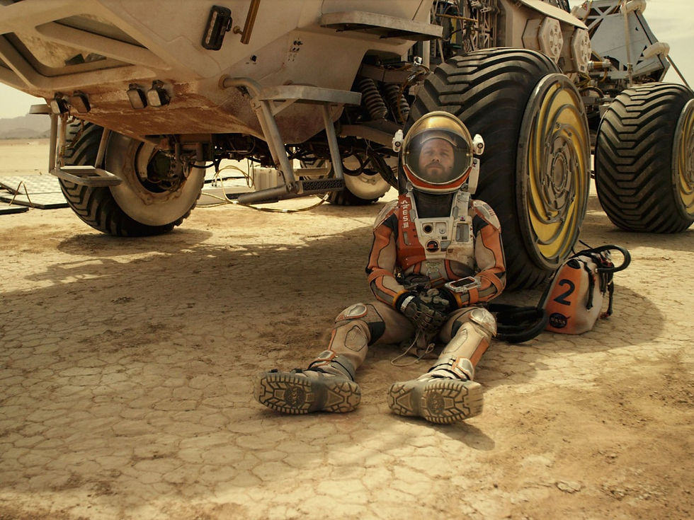Astronaut in orange suit sits by large vehicle on dry, cracked ground. Helmet rests beside, marked "2" with a NASA logo. Desert setting.