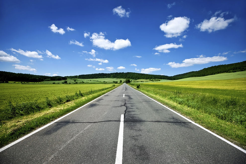 Empty rural road stretching into distance, surrounded by green fields under a clear blue sky with fluffy clouds. Peaceful and open.