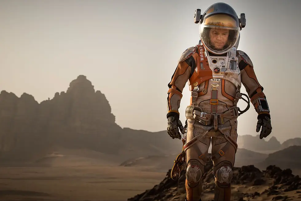 Astronaut in an orange and beige suit stands on a rocky, barren landscape with distant mountains, under a clear sky. Helmet reflects light.