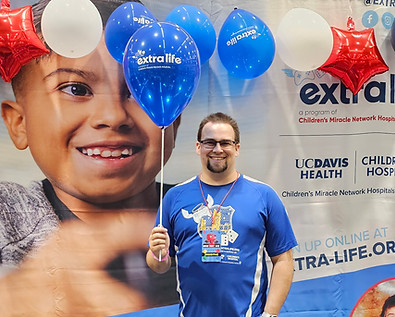 A man stands with a smile as his hand is in his pocket and the other hand is holding three balloons