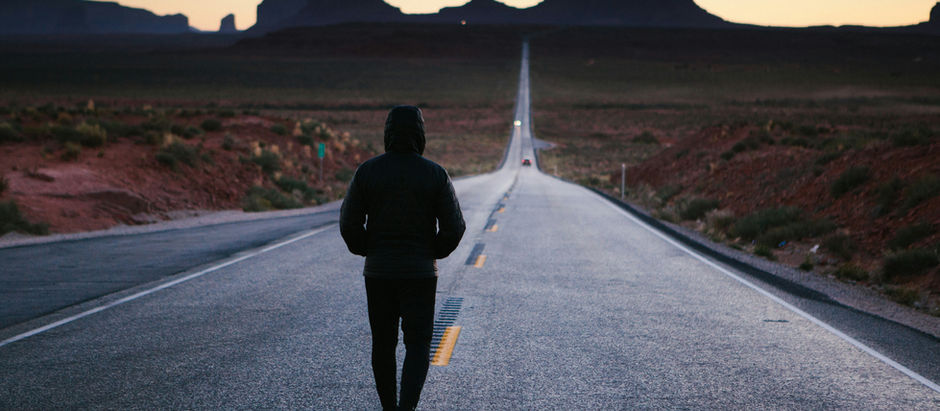 Une personne vue de dos marche au centre d'une route déserte qui file vers l'horizon, au crépuscule, avec des formations rocheuses au loin.