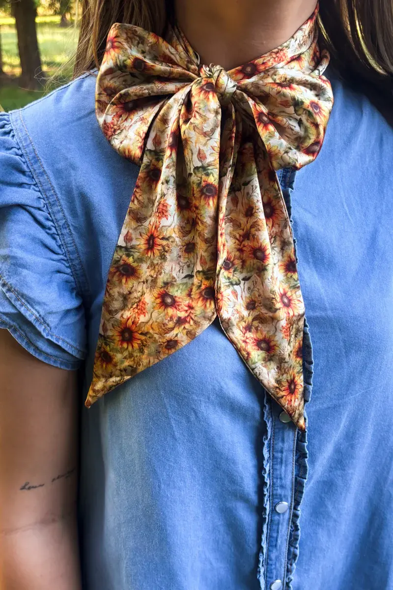 Woman wearing a vintage sunflower print western twilly scarf tied around her neck with a blue denim blouse.