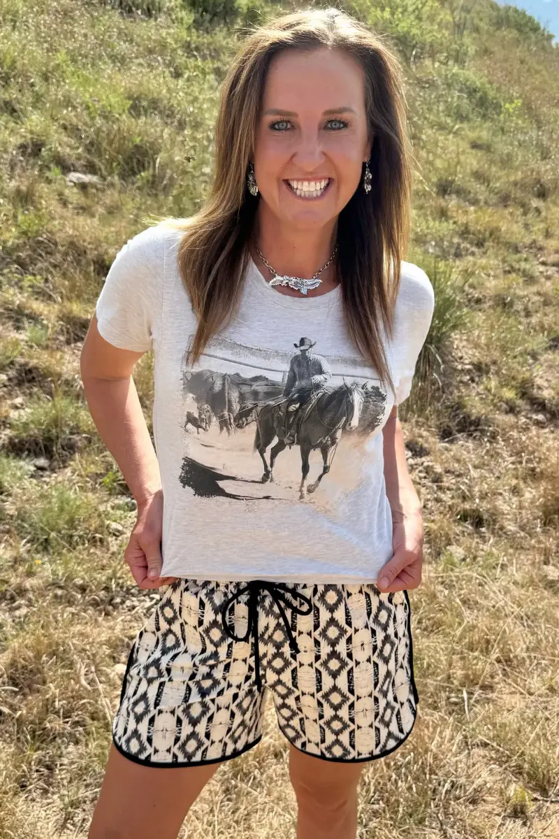 Woman wearing the To The Fire western graphic tee by Sterling Kreek paired with western print shorts in an outdoor rustic set
