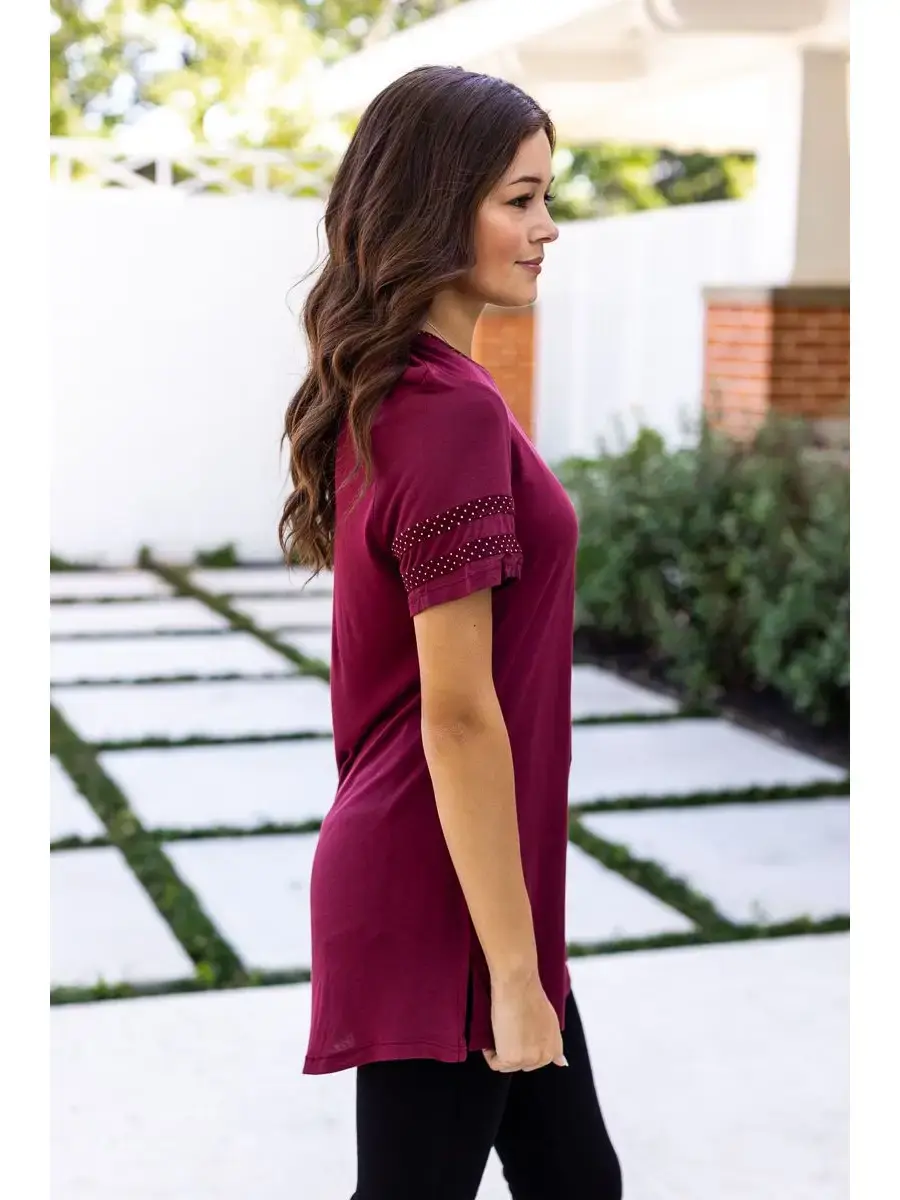 Thumbnail: "Model wearing a burgundy tunic top with polka dot detailing on the sleeves and neckline, standing outdoors."