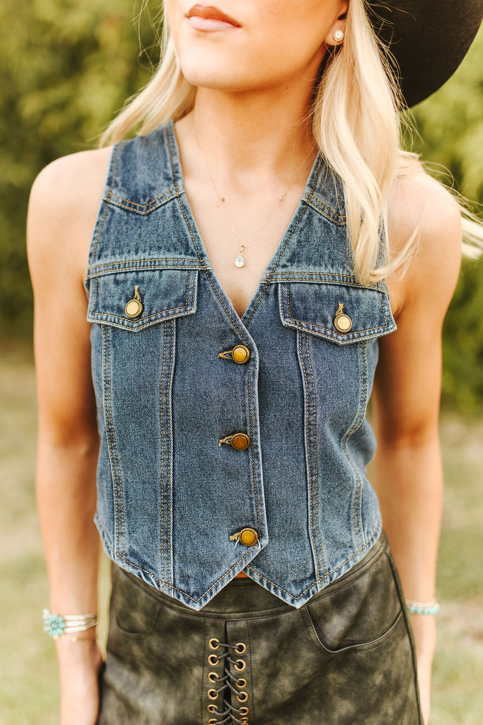 Woman wearing Lucky & Blessed mid wash button down denim vest with black western hat and western-style skirt outdoors