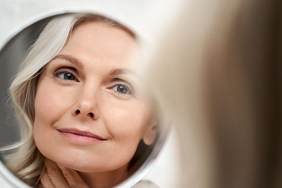 Happy 50s middle aged woman model touching face skin looking in mirror reflection. Smiling