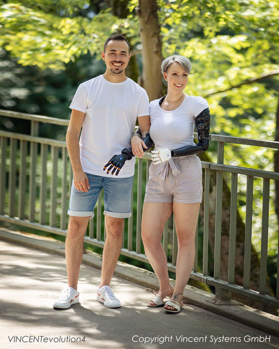 Zwei Personen stehen gemeinsam im Freien auf einem Steg, beide tragen jeweils eine Handprothese