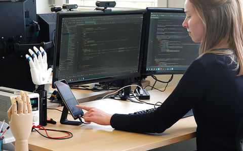 Team Elektrotechnik bei Vincent Systems bei der Arbeit: Eine Person arbeitet an einem Arbeitsplatz mit zwei Monitoren an der elektronischen Entwicklung einer Handprothese, daneben stehen Prothesenhände als Test- und Anschauungsmodelle.