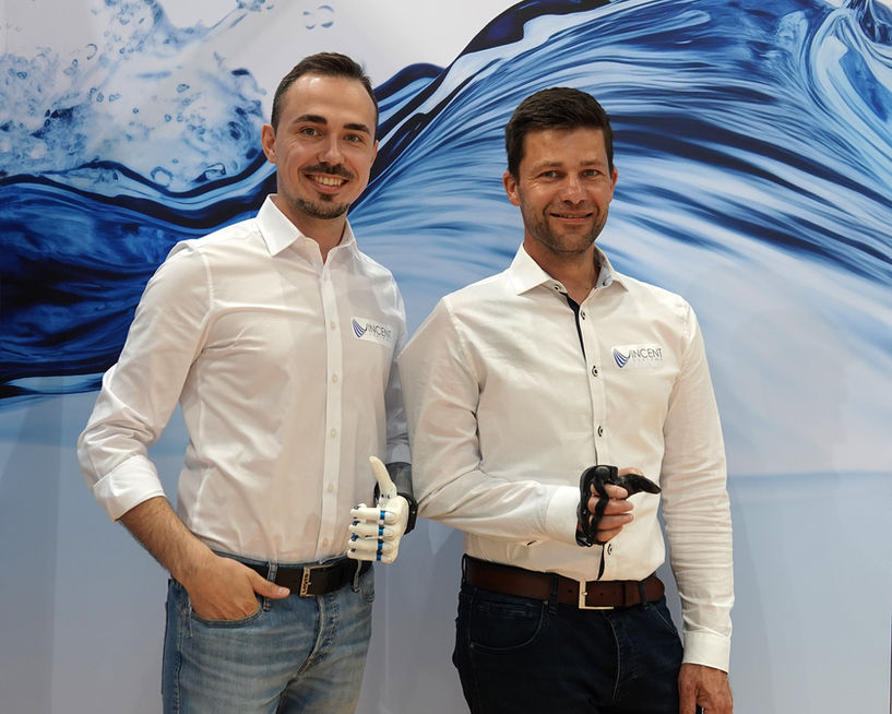 Zwei Personen auf einem Messestand: Eine Person trägt ein Exoskelett, die andere eine multiartikulierende Handprothese, präsentiert vor einer grafischen Wasserdarstellung
