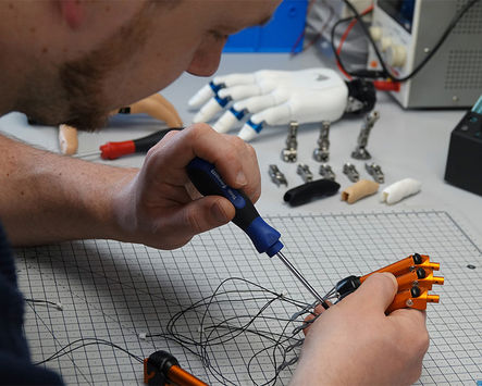 Produktion bei Vincent Systems: Eine Person verschraubt Bauteile an einer Handprothese auf einer Werkbank, mit sichtbaren Kabeln und Prothesenkomponenten.