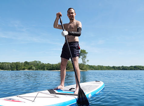 Person mit Handprothese steht auf einem Stand-up-Paddleboard auf einem See und paddelt auf ruhigem Wasser