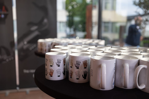 Mehrere Tassen mit Handprothesenmotiv sind geordnet auf einem runden Tisch in einem Ausstellungsbereich platziert