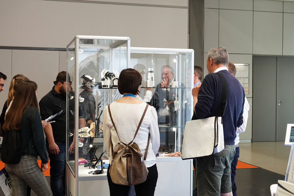 Besucherinnen und Besucher betrachten eine Glasvitrine mit Prothesen und Assistenzsystemen auf einer Messe
