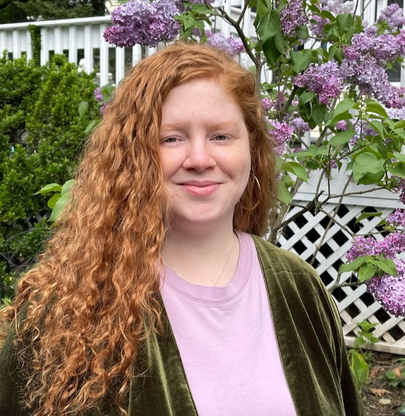 Smiling red-haired woman in a green jacket and pink shirt stands by blooming lilacs and a white fence in a garden.