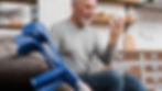 Crutches resting against a chair in the foreground, in the background a man is sitting looking at a bottle of medicine.