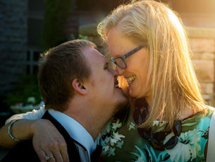 A lady and young man share an affectionate moment, touching noses and smiling, in a sunlit outdoor setting. The woman wears a floral dress and glasses.