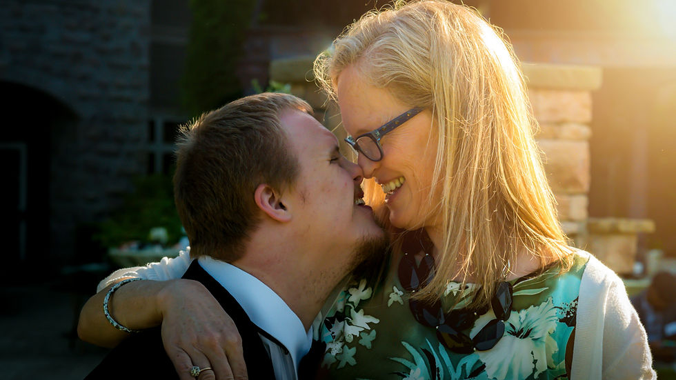 A lady and young man share an affectionate moment, touching noses and smiling, in a sunlit outdoor setting. The woman wears a floral dress and glasses.
