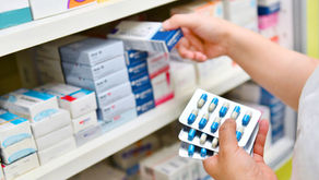 Hands selecting blue-and-white medicine boxes on pharmacy shelves while holding blister packs of blue capsules.