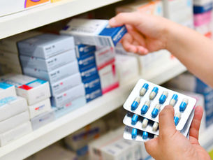 Hands selecting blue-and-white medicine boxes on pharmacy shelves while holding blister packs of blue capsules.