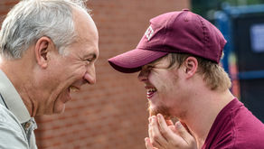 Two men smiling at each other outdoors; one wears a maroon cap and shirt, the other in a grey shirt. Brick wall and blue fence in background.